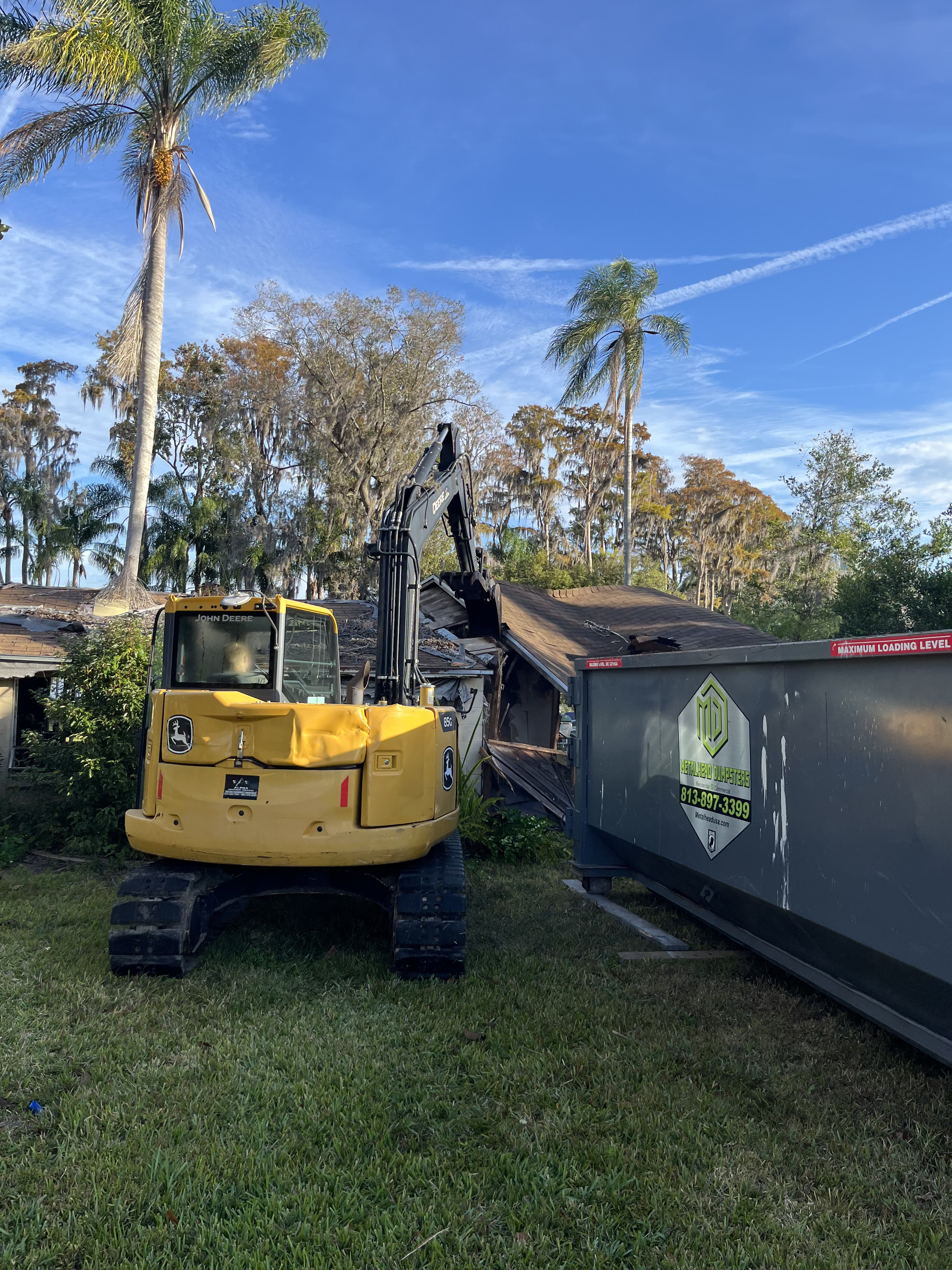 Excavator demolishing a home's roof framing next to a debris dumpster